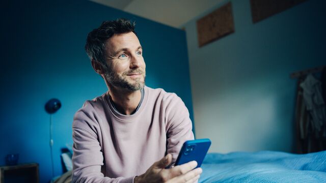 Man sitting on a bed, holding his phone and looking up - Small